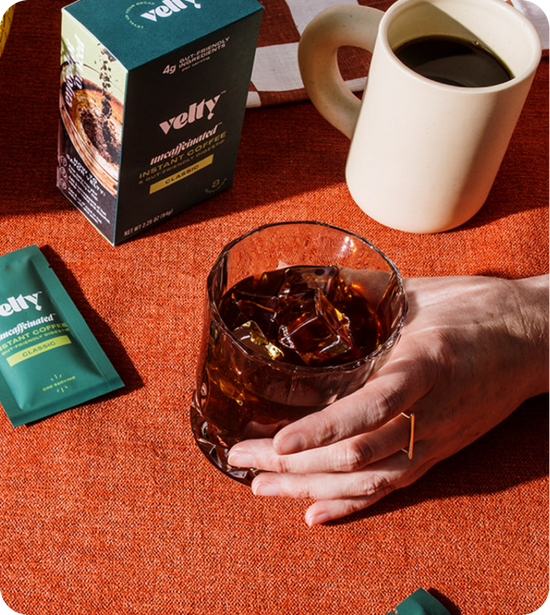 female hand holding iced coffee surrounded by Velty packet, box, and mug