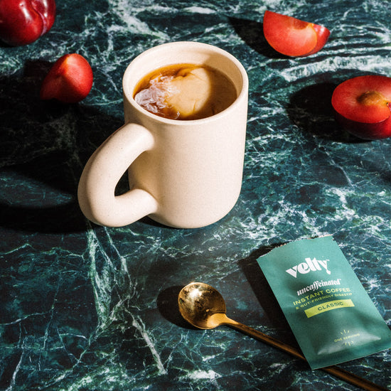 white mug filled with coffee on green table surrounded by Velty packet and red fruit