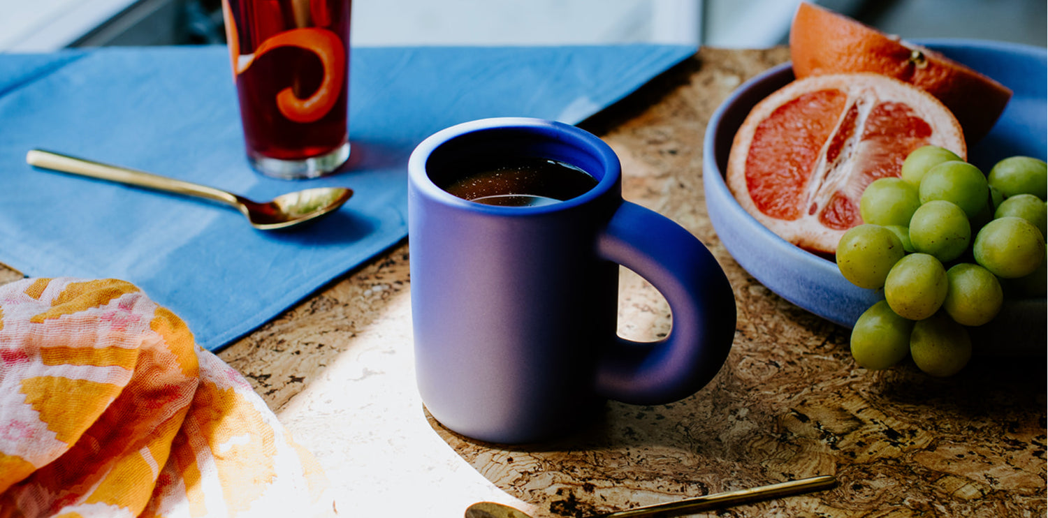 blue mug filled with coffee on table surrounded by napkins and breakfast fruits
